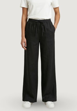 Osoba nosící volné černé kalhoty na stažení, bílé tričko s krátkým rukávem a bílé tenisky, stojící s rukama v kapsách.