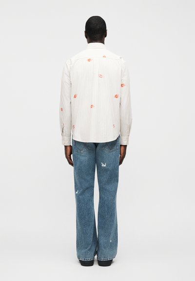 Ριγέ λευκό πουκάμισο με πορτοκαλί αποτυπώματα χειλιών στην πλάτη, συνδυασμένο με τζιν καμπάνα σε μπλε χρώμα με σκισίματα και μαύρα παπούτσια.