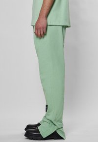 Helles grünes Baumwollmischung-Sweatpants mit geraden Beinen, umgeschlagenen Bündchen und einem Stoffetikett auf der linken Seite, kombiniert mit schwarzen klobigen Schuhen.