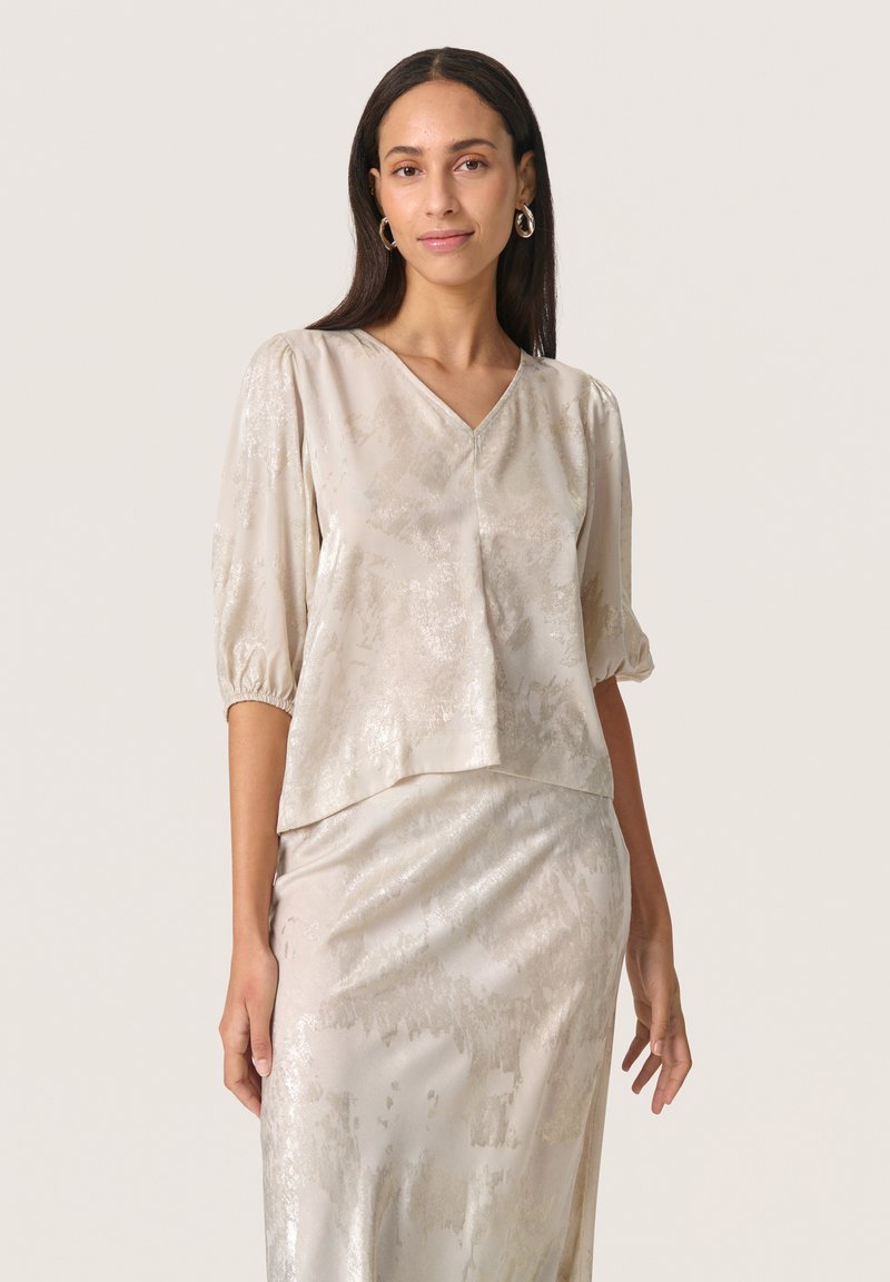 Light beige top with a V-neck and puff sleeves, made of silky fabric featuring a subtle sheen and textured pattern. Paired with a matching skirt.