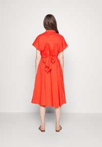 Kvinna som står vänd bort, iklädd en ljusröd midiklänning med knytbart midjeband och uppvikta korta ärmar, tillsammans med platta sandaler.