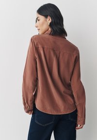 Barna, texturált hosszú ujjú ing hátsó pánttal és begombolt mandzsettával, sötét farmerrel párosítva. Tiszta, szabott sziluett.