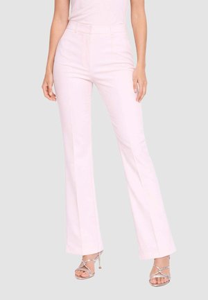 Donna che indossa pantaloni dritti vita alta color rosa chiaro e sandali argentati con tacco alto e cinturini, in piedi davanti a uno sfondo semplice.