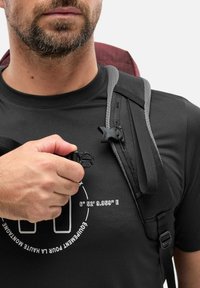 Homme avec une barbe ajustant la sangle d'un sac à dos noir sur une chemise noire avec du texte blanc et des coordonnées sur la poitrine, fond clair.