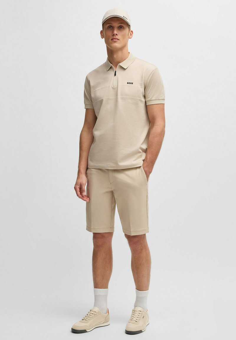 Polo beige con colletto con zip, abbinato a pantaloni corti beige. L'outfit presenta un branding discreto ed è realizzato in tessuto leggero.