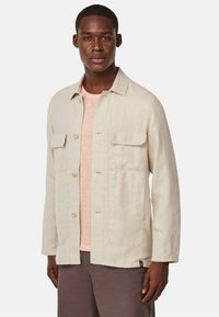 Helle beige Leinenjacke mit Kragen, zwei Fronttaschen und Knopfverschluss. Getragen über einem hellrosa T-Shirt und braunen Hosen.