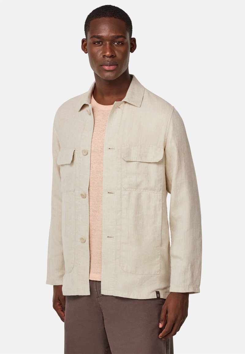 Helle beige Leinenjacke mit Kragen, zwei Fronttaschen und Knopfverschluss. Getragen über einem hellrosa T-Shirt und braunen Hosen.