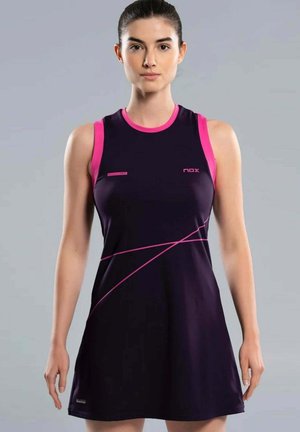 Vrouw die een mouwloze zwarte en roze sportjurk draagt met geometrische roze lijnen op de rok, staande tegen een eenvoudige grijze achtergrond.