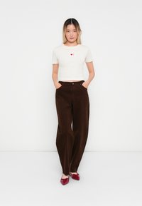 Vrouw die met handen in de zakken staat, gekleed in een witte cropped T-shirt met harticonen, een paarse wijde broek en rode puntige schoenen, tegen een witte muur.