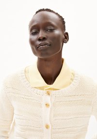 Strickjacke in Creme mit strukturiertem Muster, Rundhalsausschnitt, Knopfverschluss und einem gelben Hemd mit Kragen darunter.