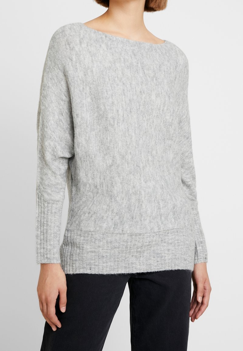 Femme portant un pull en maille douce gris clair à large encolure et un pantalon noir, debout devant un fond uni.