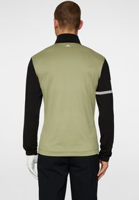 Grön och svart långärmad golftröja med en andningsbar textur, kontrasterande ärmar och en liten logotyp på bakre kragen.