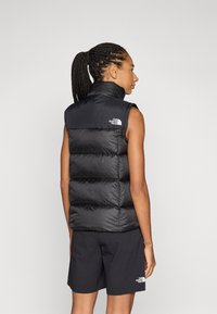 Svart pufferväst med hög krage, med vertikalt vadderade sektioner och logotyp på övre vänstra sidan. Bärs med matchande svarta shorts.