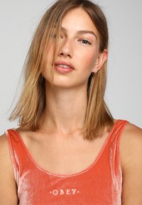 Camiseta naranja aterciopelada sin mangas con escote en forma de barco, que presenta una impresión blanca de "OBEY" centrada en el pecho. Textura suave y diseño sin mangas.