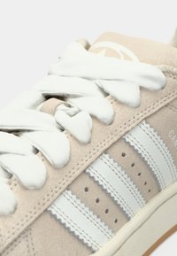 Απαλή μπεζ δερμάτινη αθλητική παπούτσια με λευκά κορδόνια και τρεις λευκές ρίγες. Διαθέτει ραμμένο σχέδιο και υφασμένη σόλα.