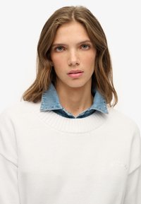 Jonge vrouw met lichtbruin golvend haar, gekleed in een witte trui over een spijkerhemd met kraag, kijkend recht vooruit tegen een witte achtergrond.