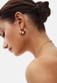 Pendiente de aro dorado con una superficie lisa y reflectante, que presenta un diseño curvado. Lleva puesto en la oreja izquierda, el tono de piel es neutro.