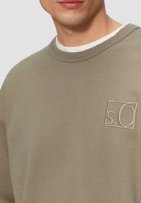 Man in een beige rondhalstrui met een klein geborduurd "s.O" logo op de borst, gedragen over een witte rondhalshem. 
