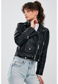 Zwarte leren bikerjack met zilveren ritsen, drukknoopsluitingen en een ceintuur in de taille, gestyled met lichtblauwe high-waisted jeans.