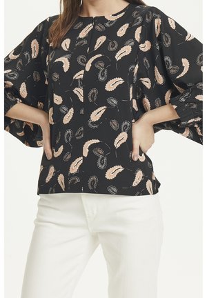 Kvinde i sort bluse med lyserøde og beige bladmønstre, puffede ærmer, kombineret med hvide bukser, hænderne på hofterne.