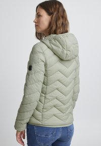 Veste rembourrée légère verte avec capuche, présentant un motif matelassé en chevrons et un emblème noir sur la manche gauche. Coupe décontractée.