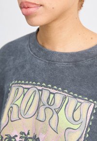 Grijze geribde sweatshirt met een kleurrafische 'ROXY' afbeelding met palmboomontwerp en een gekartelde rand langs de halslijn.