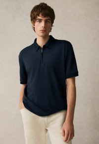 Mörkblå polo med dragkedja i kragen, kortärmad och med texturerade kanter, matchad med ljusa byxor. Slät tyg med avslappnad passform.