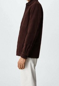 Giacca di velluto a coste marrone con chiusura a bottoni, taschino sul petto e maniche lunghe. Indossata con pantaloni di colore chiaro.
