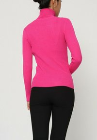 Col roulé ajusté en rose vif, fabriqué en tissu extensible. Manches longues et ourlet légèrement arrondi à l'arrière. Porté avec un pantalon noir.