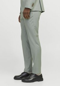 Světle zelené kalhoty formovaného střihu vyrobené z hladké tkaniny, spárované s černými koženými botami a světle barevnými ponožkami. Slim fit design se bočními švy.