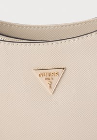 Beżowa teksturowana torba na ramię z trójkątną złotą plakietką z logo "GUESS" i ikoną, podkreślona kontrastującym ciemnym zamkiem błyskawicznym.