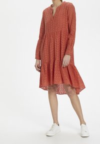 Šaty v rezavé barvě s dlouhými rukávy, vyšívanými výstřihovými detaily, výstřihem do V a vrstveným spodním lemem. Doplněno bílými teniskami.