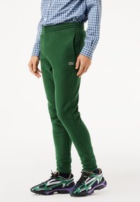 Gröna bomullsoveraller med elastisk midja och sidofickor, med en vit Lacoste-logotyp på vänster lår. Bärs med flerfärgade sneakers.