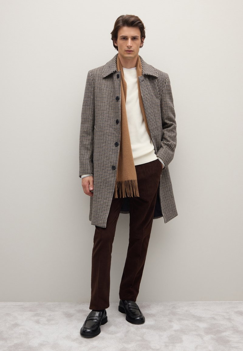 Houndstooth patroon overjas in bruin en zwart, crèmekleurige trui, bruine corduroy broek, bruin franjesjaal en zwarte loafers.