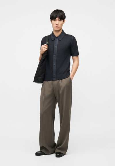 Νεαρός άνδρας με μαύρο κοντομάνικο πουκάμισο με κουμπιά, φαρδιά καφέ παντελόνια, μαύρα παπούτσια, κρατώντας μαύρη τσάντα ώμου, στέκεται σε λευκό φόντο.