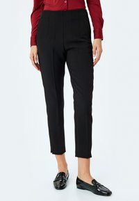 Pantalons noirs ajustés avec des détails plissés, coupe slim et longueur raccourcie. Associés à des mocassins noirs, portés avec une chemise boutonnée bordeaux.