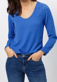 Blaue Langarmbluse mit tiefem V-Ausschnitt und glatter Textur, kombiniert mit dunkelblauen Jeans im klassischen Fünf-Taschen-Design.