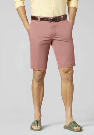 Mann trägt rosafarbene Shorts, hellgelbes Hemd, braunen Gürtel und grüne karierte Sandalen, steht mit den Händen in den Taschen.