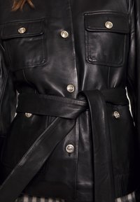Schwarze Lederjacke mit zwei Fronttaschen, silbernen Knopfdetails und einem passenden Gürtel, mit einer glatten Textur und tailliertem Design.