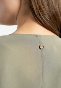 Oliwkowa bluzka o gładkiej teksturze, prostokątnym dekolcie i ozdobnym okrągłym guziku z napisem "CLICK" na plecach.
