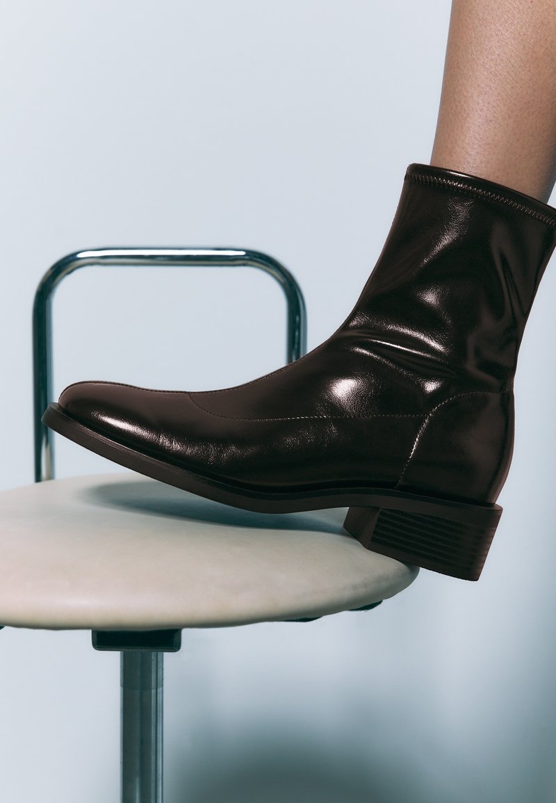 Bottine en cuir marron avec une finition lisse et brillante, un bout arrondi et un petit talon empilé. Placée sur un tabouret de couleur claire.