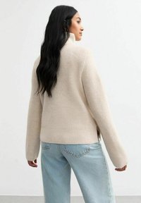 Pull en mailles beiges avec col montant, fentes latérales à l'ourlet, porté avec un jean bleu clair. Texture douce, coupe décontractée et manches longues.