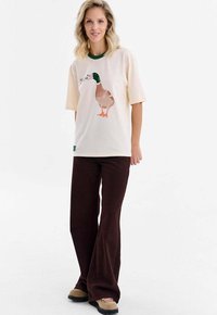 Krémové tričko se zeleným límcem s potiskem kachny a volným střihem. Kombinováno s hnědými kalhotami širokého střihu a béžovou obuví.