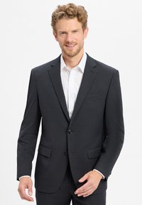 Giacca da completo da uomo grigio scuro, con chiusura a due bottoni, rever a notches e due tasche frontali. Indossata sopra una camicia bianca. Tessuto di texture liscia.