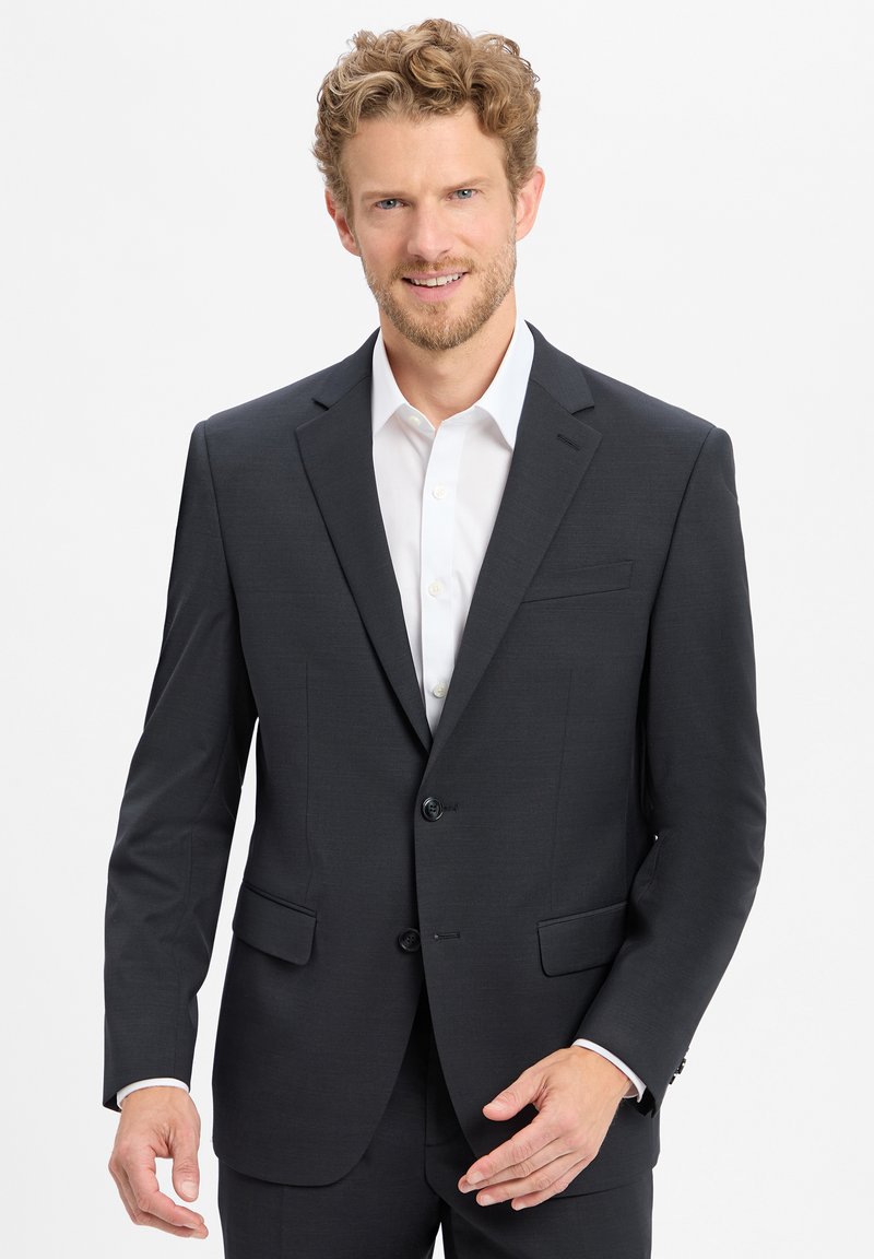 Giacca da completo da uomo grigio scuro, con chiusura a due bottoni, rever a notches e due tasche frontali. Indossata sopra una camicia bianca. Tessuto di texture liscia.