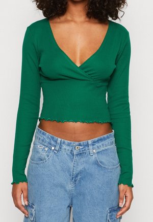 Mujer con un top corto acanalado de manga larga verde con bajo ondulado y pantalones cargo de mezclilla azul claro de tiro alto.