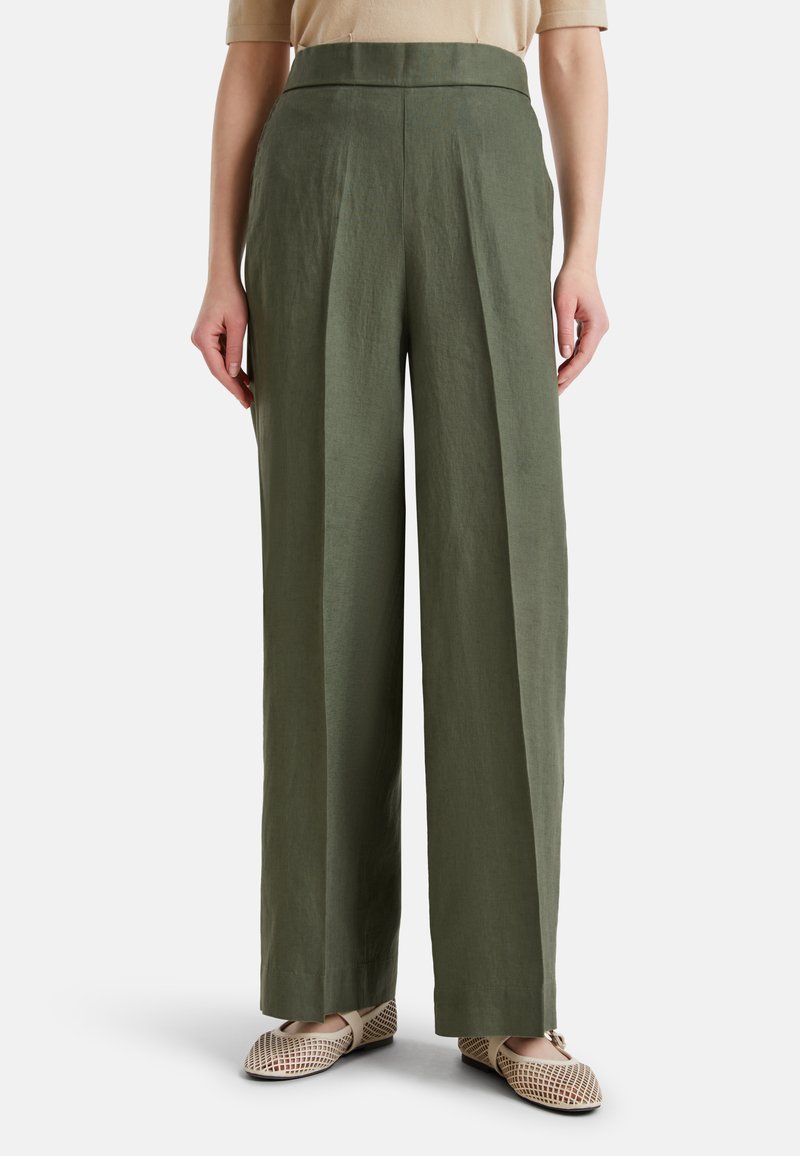 Mujer con pantalones de talle alto y pierna ancha en verde oliva, combinados con bailarinas beige tejidas, de pie frente a un fondo liso.