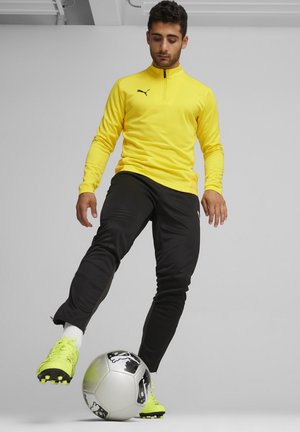Geel sportieve longsleeve shirt met een ritssluiting bij de kraag, gecombineerd met zwarte broek en gele voetbalschoenen, geplaatst bovenop een voetbal.