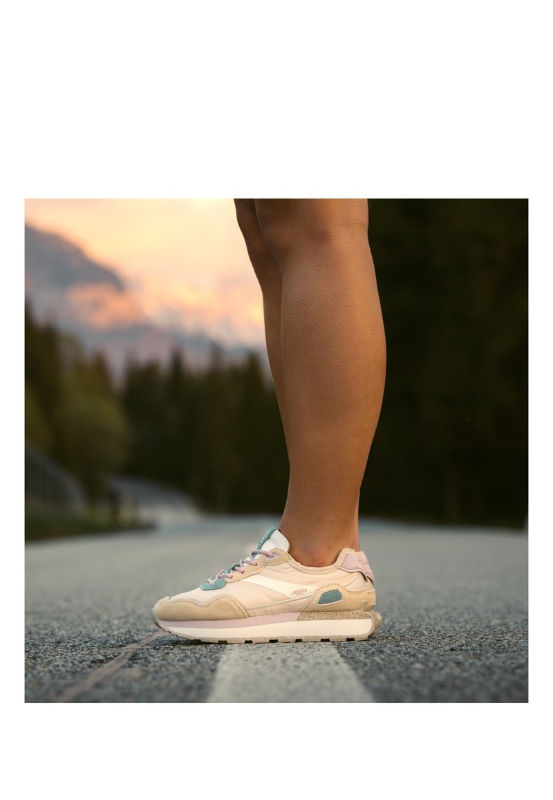 Jambes portant des baskets beige et blanches debout sur une route asphaltée avec des arbres flous et un ciel au coucher du soleil en arrière-plan.