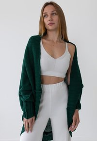 Długi zielony dzianinowy kardigan na białej prążkowanej bluzce crop top i białych spodniach z wysokim stanem. Gładkie tekstury o luźnym kroju i otwartych przodzie.